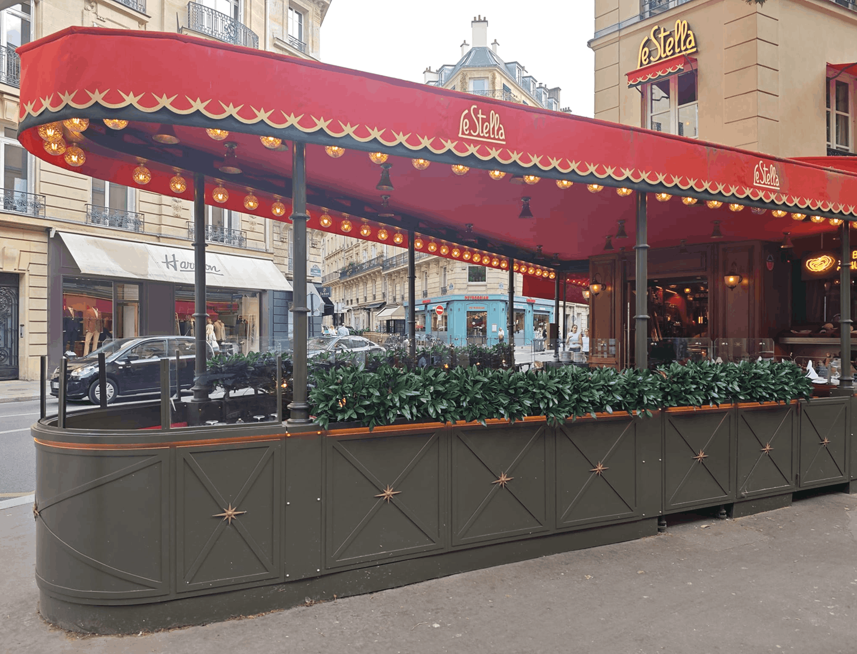Terrasse extérieure sur mesure du restaurant Le Stella Terrasse extérieure sur mesure réalisée pour le restaurant Le Stella avec paravents vitrés télescopiques, jardinières intégrées et éclairage à ampoules filament.